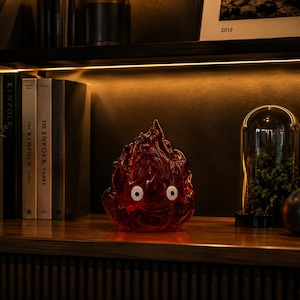 May include: A translucent red flame-shaped figurine with two white circular eyes featuring black pupils. The object has a textured, undulating surface that mimics the appearance of flickering fire, standing as a decorative piece on a wooden shelf.