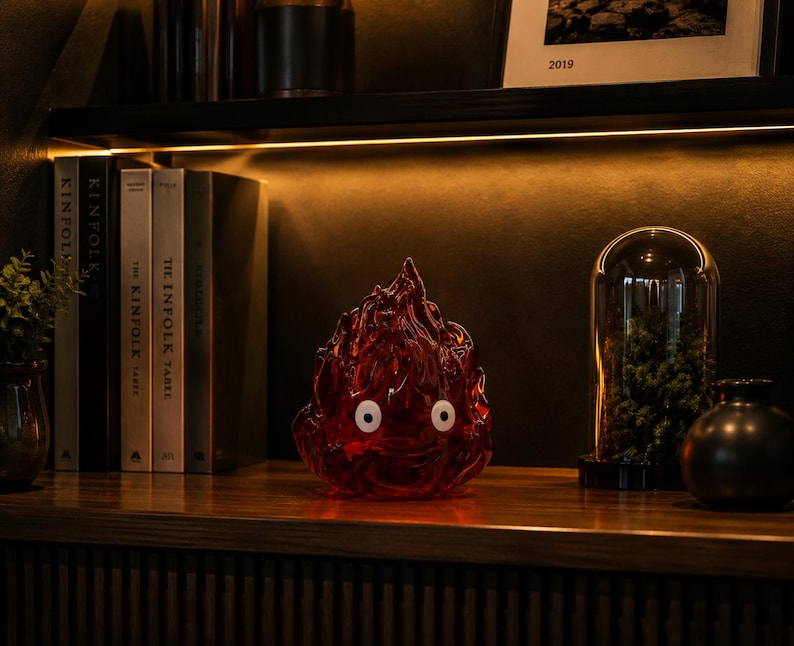 May include: A translucent red flame-shaped figurine with two white circular eyes featuring black pupils. The object has a textured, undulating surface that mimics the appearance of flickering fire, standing as a decorative piece on a wooden shelf.