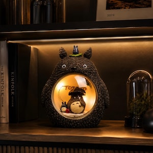 K&ouml;nnte beinhalten: Eine dunkelgraue, texturierte Lampe in Totoro-Form mit einem kreisf&ouml;rmigen Ausschnitt in der Mitte. Im beleuchteten Bereich h&auml;lt eine kleine Totoro-Figur einen schwarzen Regenschirm neben einer winzigen blauen Figur. Oben auf der Lampe sitzt eine kleine graue Figur.