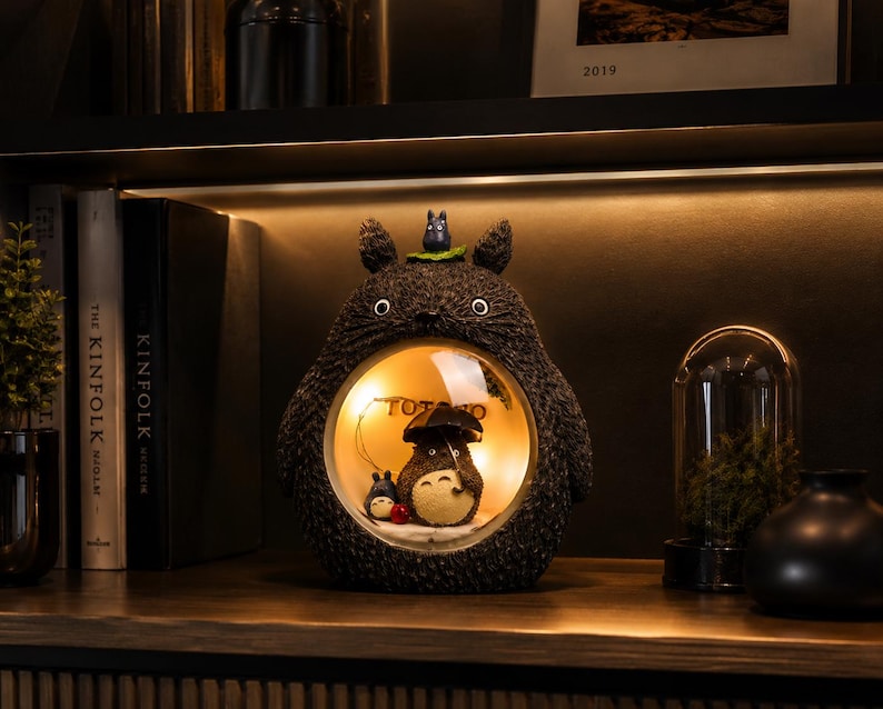 K&ouml;nnte beinhalten: Eine dunkelgraue, texturierte Lampe in Totoro-Form mit einem kreisf&ouml;rmigen Ausschnitt in der Mitte. Im beleuchteten Bereich h&auml;lt eine kleine Totoro-Figur einen schwarzen Regenschirm neben einer winzigen blauen Figur. Oben auf der Lampe sitzt eine kleine graue Figur.