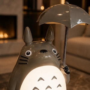May include: A grey ceramic lamp in the shape of a cartoon character, holding a grey umbrella. The lamp has a glowing, rounded base with three curved cutouts. The character has large eyes and pointed ears. The background is blurred, with a fireplace and furniture.