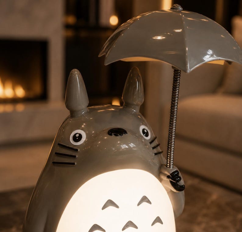May include: A grey ceramic lamp in the shape of a cartoon character, holding a grey umbrella. The lamp has a glowing, rounded base with three curved cutouts. The character has large eyes and pointed ears. The background is blurred, with a fireplace and furniture.