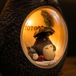 K&ouml;nnte beinhalten: Eine dunkelbraune, texturierte, eif&ouml;rmige Lampe mit einem kreisf&ouml;rmigen Ausschnitt, der eine Szene mit zwei grauen, rundlichen Kreaturen beleuchtet. Eine Kreatur h&auml;lt einen schwarzen Regenschirm und der Text TOTO ist an der Innenwand zu sehen.