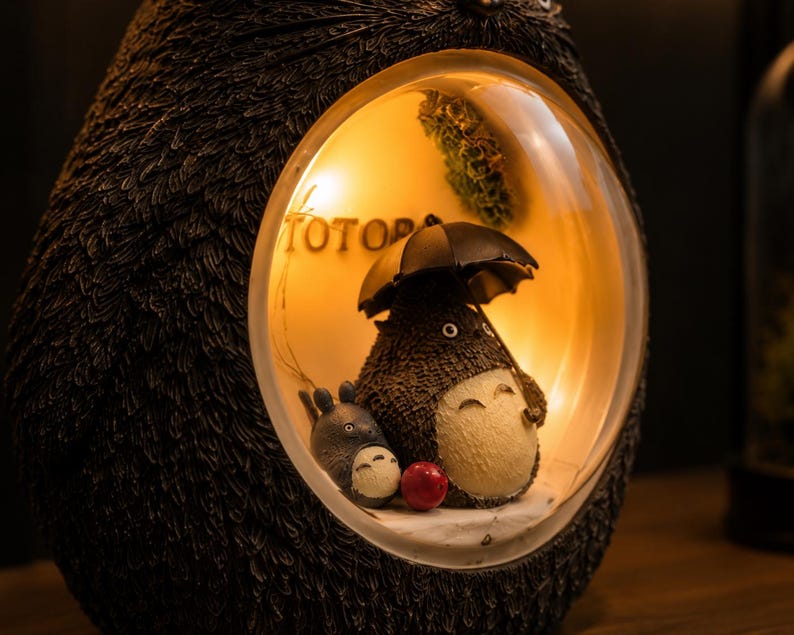 K&ouml;nnte beinhalten: Eine dunkelbraune, texturierte, eif&ouml;rmige Lampe mit einem kreisf&ouml;rmigen Ausschnitt, der eine Szene mit zwei grauen, rundlichen Kreaturen beleuchtet. Eine Kreatur h&auml;lt einen schwarzen Regenschirm und der Text TOTO ist an der Innenwand zu sehen.