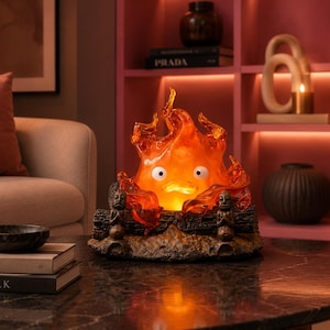 Op de afbeelding: Decoratieve lamp met een oranje vlamontwerp en een cartoongezicht. De lamp staat op een voet met gebeeldhouwde figuren en een log-achtig uiterlijk. De lamp staat op een donkere marmeren tafel.
