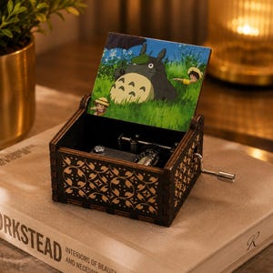 May include: A dark wooden music box with a hand crank on the side. The lid features a colorful illustration of a large grey creature with a leaf on its head, holding an umbrella in a grassy field with two small figures. The box has intricate patterns.