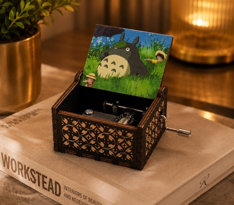May include: A dark wooden music box with a hand crank on the side. The lid features a colorful illustration of a large grey creature with a leaf on its head, holding an umbrella in a grassy field with two small figures. The box has intricate patterns.