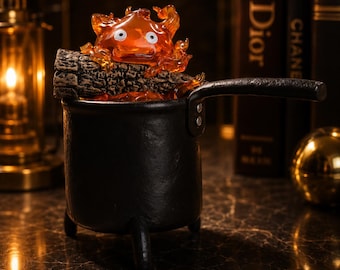 May include: A black metal pot with three legs and a side handle, containing a textured brown log and a translucent orange flame character with two white eyes. The flame figure is positioned on top of the log, which rests inside the black pot.