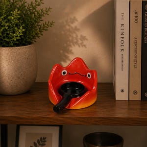 May include: A bright red ceramic flame-shaped holder with a stylised face featuring two white eyes and a thin black mouth. A small black frying pan with a long handle is positioned in the centre of the red holder, creating a contrasting visual.