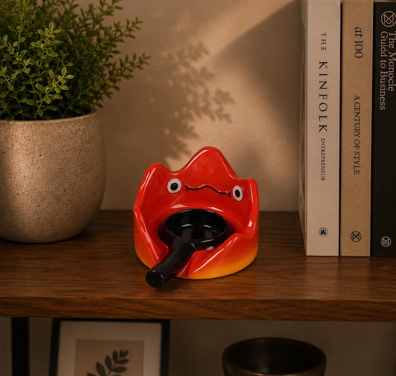 May include: A bright red ceramic flame-shaped holder with a stylised face featuring two white eyes and a thin black mouth. A small black frying pan with a long handle is positioned in the centre of the red holder, creating a contrasting visual.
