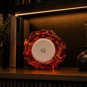 May include: A circular, amber-colored glass light fixture with a wavy, organic edge. The center features a flat, white circular panel with a black button in the middle labeled ON/OFF. The glass has a translucent, rippled texture throughout.