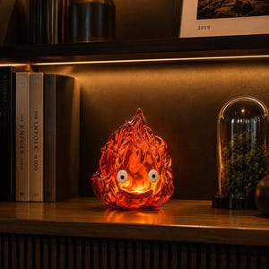 May include: A translucent orange and red flame-shaped lamp with a smooth, sculpted surface. The lamp features two white circular eyes and a wide, curved mouth, emitting a warm internal glow that illuminates its detailed, fluid-like texture.