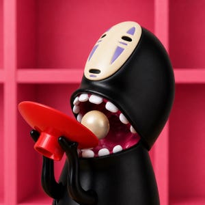 May include: A black figurine of a character with a white face and purple markings, holding a red plate. The character's mouth is open, revealing white teeth and a pearl-like sphere. The background is a pink shelf.