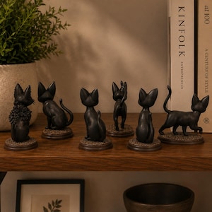 May include: A collection of six small black cat figurines displayed on a wooden shelf. Each cat is depicted in a different pose, including sitting, standing, and walking, and is mounted on a circular base with a textured, brown, ground-like finish.