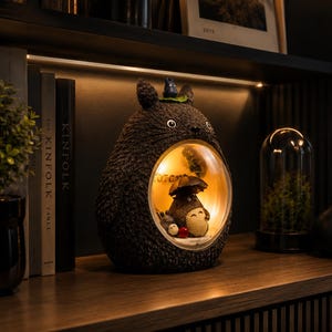 K&ouml;nnte beinhalten: Eine dunkelbraune, texturierte Lampe in Totoro-Form mit einem kreisf&ouml;rmigen Ausschnitt, der eine beleuchtete Innenszene enth&uuml;llt. Im Inneren h&auml;lt eine kleine Totoro-Figur einen Regenschirm, begleitet von zwei winzigen Ru&szlig;m&auml;nnchen. Obenauf sitzt eine kleine blaue Figur.