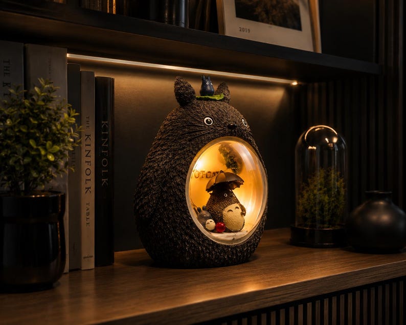 K&ouml;nnte beinhalten: Eine dunkelbraune, texturierte Lampe in Totoro-Form mit einem kreisf&ouml;rmigen Ausschnitt, der eine beleuchtete Innenszene enth&uuml;llt. Im Inneren h&auml;lt eine kleine Totoro-Figur einen Regenschirm, begleitet von zwei winzigen Ru&szlig;m&auml;nnchen. Obenauf sitzt eine kleine blaue Figur.
