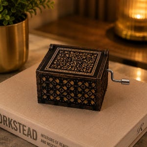 May include: A square, black wooden music box with a hand crank on the side. The lid and sides feature intricate, laser-cut floral and geometric patterns that reveal a light-colored interior layer beneath the dark exterior surface.