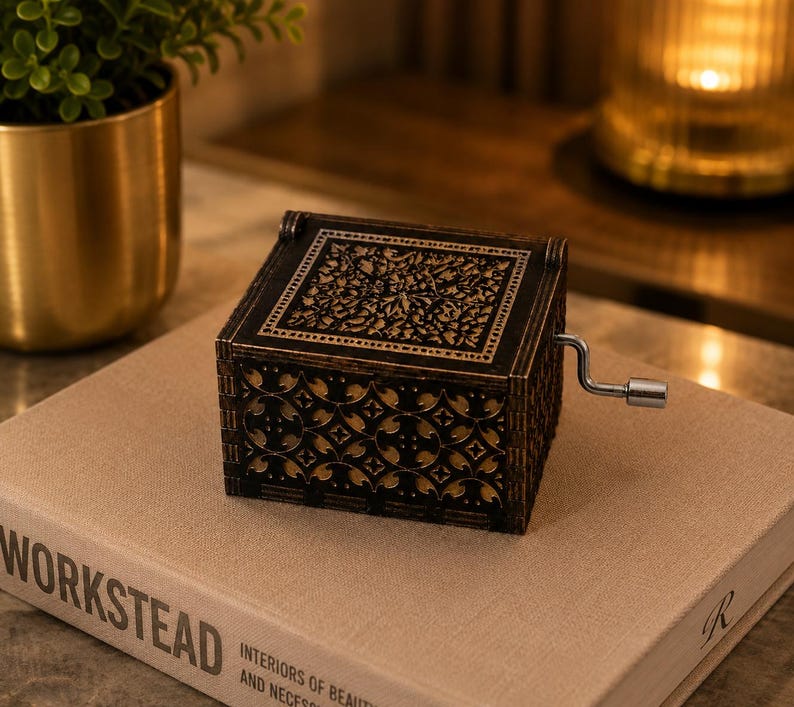 May include: A square, black wooden music box with a hand crank on the side. The lid and sides feature intricate, laser-cut floral and geometric patterns that reveal a light-colored interior layer beneath the dark exterior surface.