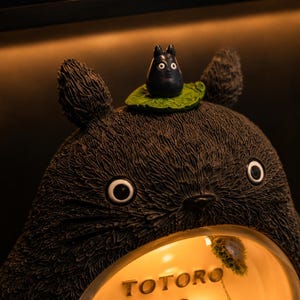 K&ouml;nnte beinhalten: Eine dunkelgraue, texturierte Totoro-Figur mit spitzen Ohren und gro&szlig;en Augen. Eine kleine schwarze Kreatur sitzt auf einem gr&uuml;nen Blatt auf dem Kopf. Der Bauch zeigt ein kreisf&ouml;rmiges, beleuchtetes Fenster mit dem Schriftzug TOTORO.