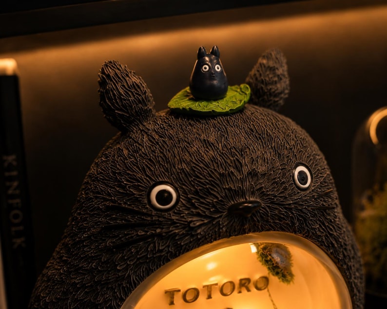 K&ouml;nnte beinhalten: Eine dunkelgraue, texturierte Totoro-Figur mit spitzen Ohren und gro&szlig;en Augen. Eine kleine schwarze Kreatur sitzt auf einem gr&uuml;nen Blatt auf dem Kopf. Der Bauch zeigt ein kreisf&ouml;rmiges, beleuchtetes Fenster mit dem Schriftzug TOTORO.