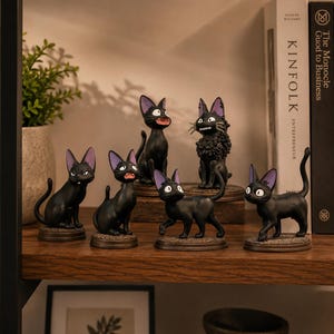 May include: A collection of six black cat figurines with purple-lined ears, each standing on a circular base. The cats are depicted in various poses, including sitting, standing, and one with a textured, curly coat, all featuring large eyes.