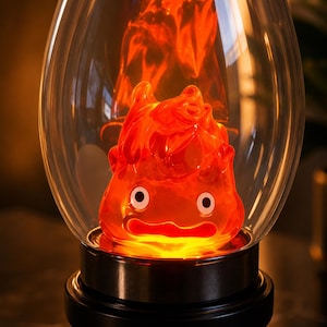 May include: A decorative lamp featuring a translucent orange flame-shaped figure with two white circular eyes and a wide mouth, enclosed within a clear glass dome. The figure sits on a black circular base with a small black knob on the side.