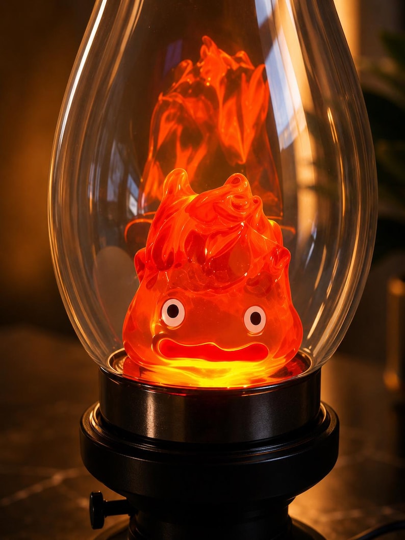 May include: A decorative lamp featuring a translucent orange flame-shaped figure with two white circular eyes and a wide mouth, enclosed within a clear glass dome. The figure sits on a black circular base with a small black knob on the side.