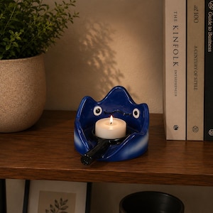 May include: A glossy blue ceramic candle holder shaped like a stylized creature with two white eyes and a black handle extending from the front. A small white tea light candle with a lit flame sits in the center of the blue ceramic vessel.