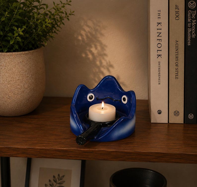 May include: A glossy blue ceramic candle holder shaped like a stylized creature with two white eyes and a black handle extending from the front. A small white tea light candle with a lit flame sits in the center of the blue ceramic vessel.