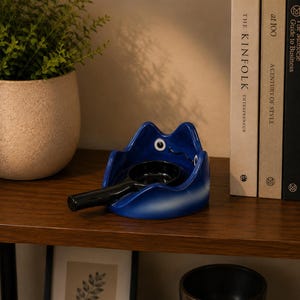 May include: A glossy blue ceramic frog-shaped ashtray with large white and black eyes. The ashtray features a deep circular basin and a long, black cylindrical handle extending from the front, designed to hold a cigarette or similar item securely.