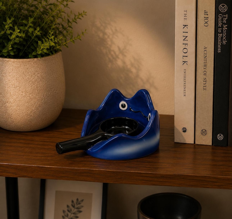 May include: A glossy blue ceramic frog-shaped ashtray with large white and black eyes. The ashtray features a deep circular basin and a long, black cylindrical handle extending from the front, designed to hold a cigarette or similar item securely.