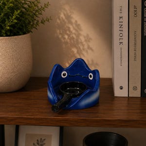 May include: A glossy blue ceramic tray shaped like a stylized creature with two white eyes and a black mouth. A small, round, black ceramic bowl with a handle is positioned in the center of the tray, creating a functional holder for small items.