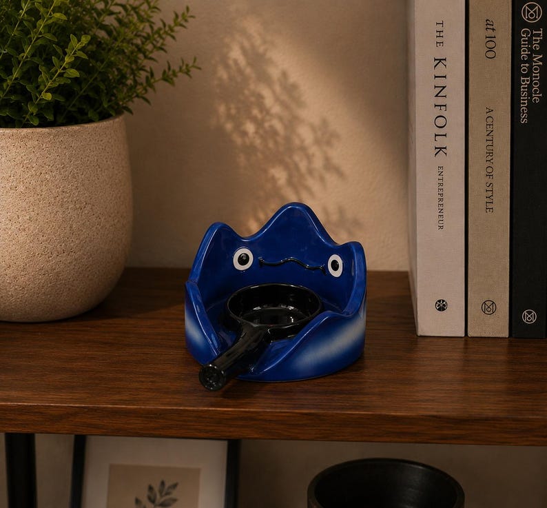 May include: A glossy blue ceramic tray shaped like a stylized creature with two white eyes and a black mouth. A small, round, black ceramic bowl with a handle is positioned in the center of the tray, creating a functional holder for small items.