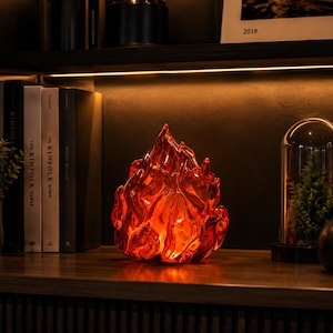 May include: A translucent, flame-shaped glass sculpture in vibrant shades of red and orange. The object features a textured, undulating surface that mimics the appearance of flickering fire, with a glowing internal light source.