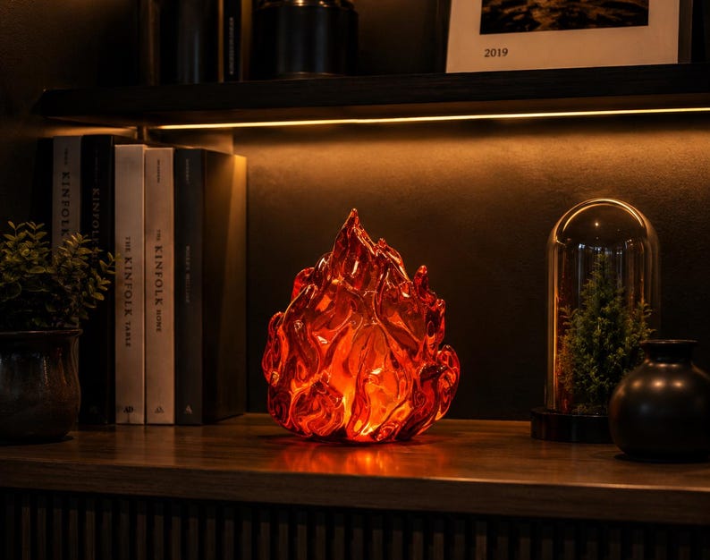 May include: A translucent, flame-shaped glass sculpture in vibrant shades of red and orange. The object features a textured, undulating surface that mimics the appearance of flickering fire, with a glowing internal light source.