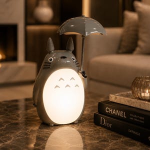 May include: A grey Totoro-shaped lamp holding a grey umbrella. The lamp has a glowing belly and is on a marble table. Books and a candle holder are in the background. The lamp is a decorative item.