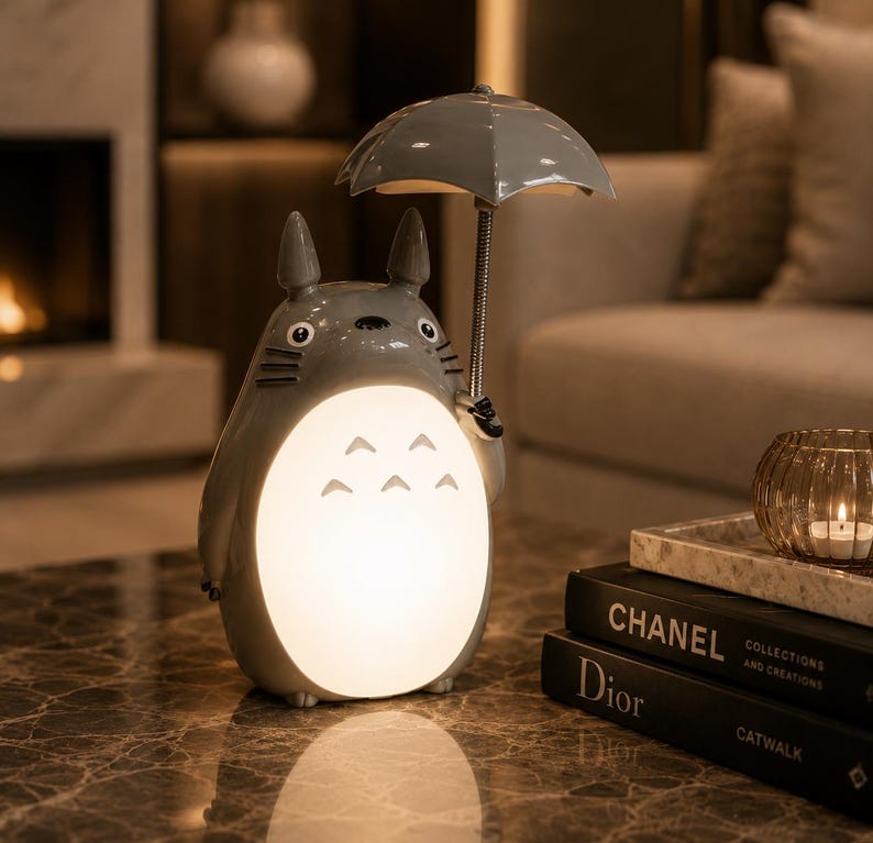 May include: A grey Totoro-shaped lamp holding a grey umbrella. The lamp has a glowing belly and is on a marble table. Books and a candle holder are in the background. The lamp is a decorative item.