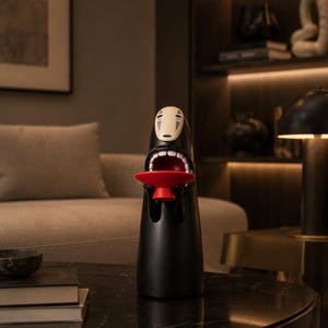 May include: A black decorative figurine with a white face and open mouth, holding a small red tray. The figurine is on a black marble table, with a neutral-toned sofa and artwork in the background.