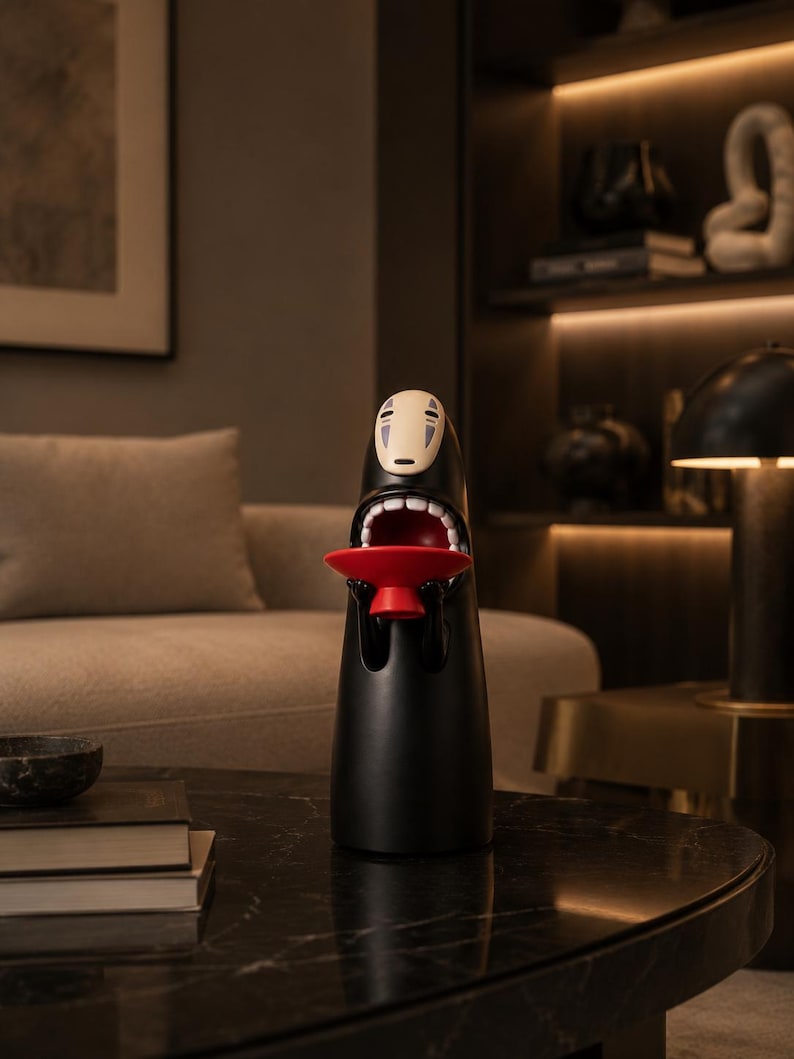 May include: A black decorative figurine with a white face and open mouth, holding a small red tray. The figurine is on a black marble table, with a neutral-toned sofa and artwork in the background.