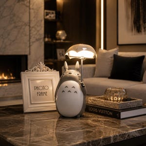 May include: A grey and white Totoro-shaped lamp holding a clear umbrella-shaped light. A white photo frame with the words "PHOTO FRAME" is to the left. A decorative tray with a glass candle holder and a book with "LOUIS VUITTON" on the cover is on the right.