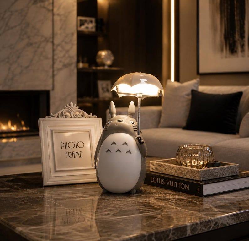 May include: A grey and white Totoro-shaped lamp holding a clear umbrella-shaped light. A white photo frame with the words "PHOTO FRAME" is to the left. A decorative tray with a glass candle holder and a book with "LOUIS VUITTON" on the cover is on the right.