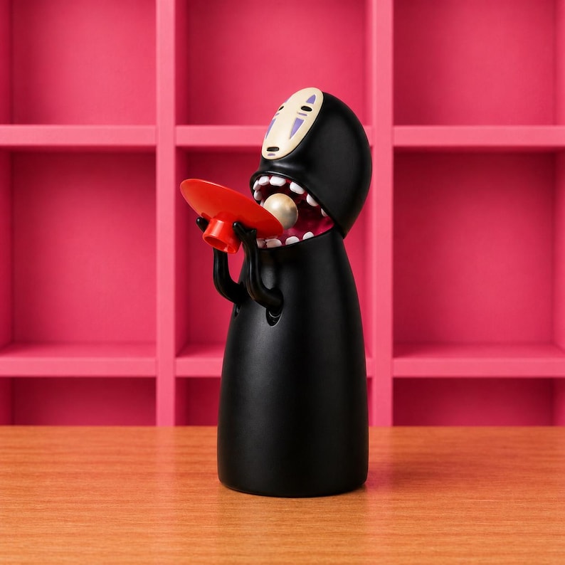 May include: A black figurine of a character from Spirited Away, holding a red plate and a pearl. The figure has a white mask with purple markings and an open mouth. The background is a pink shelf.