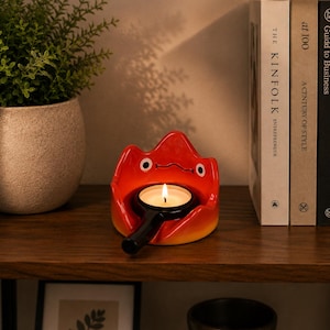 May include: A ceramic candle holder shaped like a stylised red and orange flame character with two white eyes and a simple black line for a mouth. The holder features a central circular opening containing a small black metal tea light candle holder.