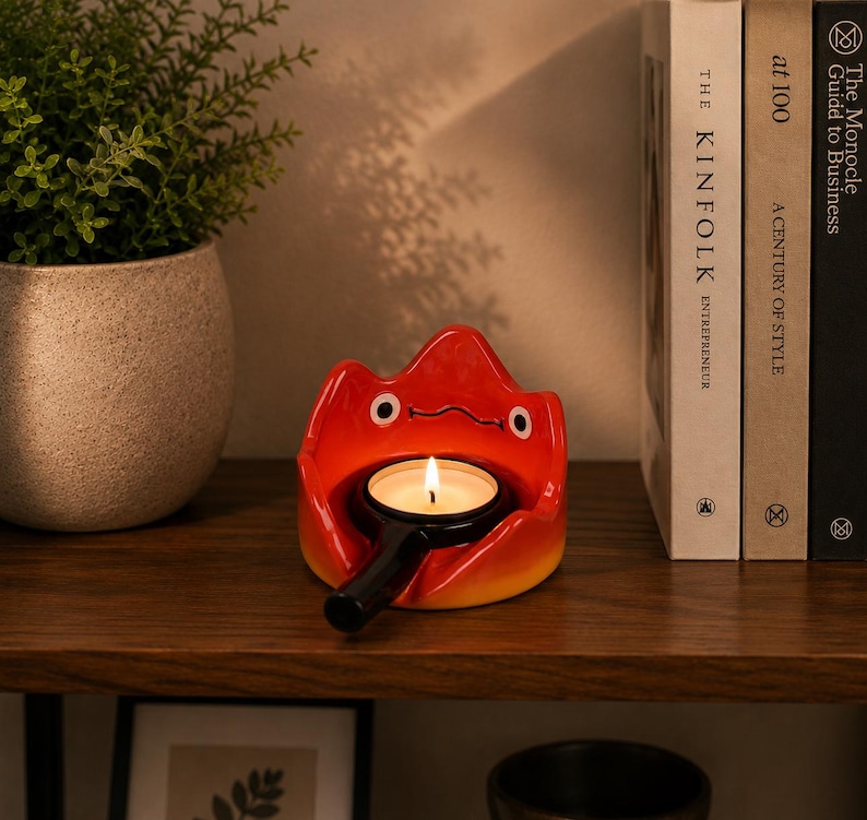 May include: A ceramic candle holder shaped like a stylised red and orange flame character with two white eyes and a simple black line for a mouth. The holder features a central circular opening containing a small black metal tea light candle holder.