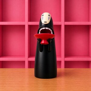 May include: A black figurine of a character from Spirited Away, holding a red bowl. The figure has a white mask-like face with purple markings and an open mouth. The background features a pink shelf unit and a wooden surface.