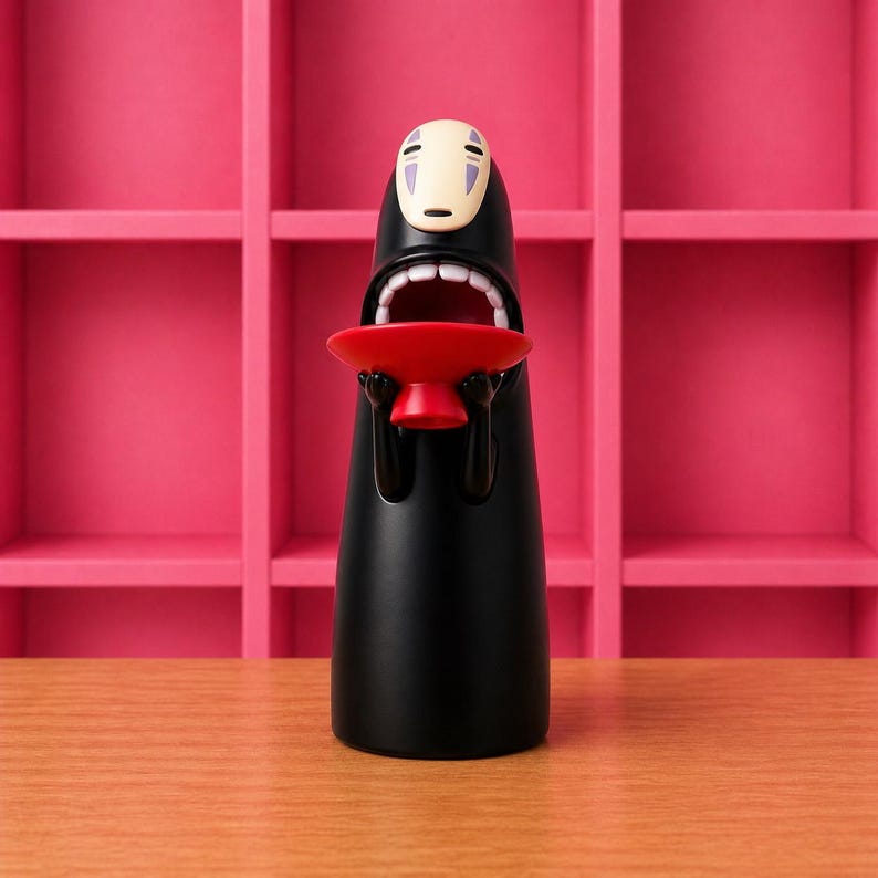 May include: A black figurine of a character from Spirited Away, holding a red bowl. The figure has a white mask-like face with purple markings and an open mouth. The background features a pink shelf unit and a wooden surface.
