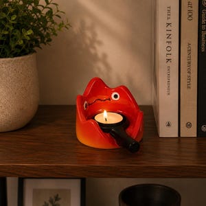 May include: A bright red and orange ceramic candle holder shaped like a stylised flame character with a black handle extending from the front. The holder contains a small, lit white tea light candle with a visible yellow flame and a black wick.