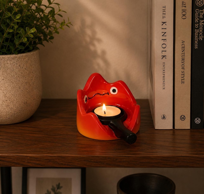 May include: A bright red and orange ceramic candle holder shaped like a stylised flame character with a black handle extending from the front. The holder contains a small, lit white tea light candle with a visible yellow flame and a black wick.