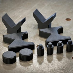 May include: A set of black metal tools, including two V-shaped pieces, two pieces with a curved top, two round pieces, and three cylindrical pieces with screw tops. The tools are arranged on a textured, gray surface.