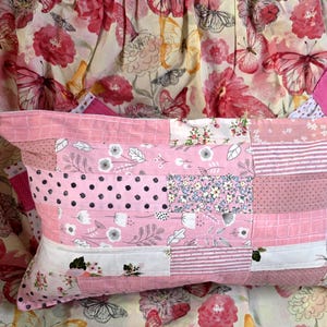 May include: A rectangular patchwork pillow in shades of pink and white. The pillow features various fabric patterns, including polka dots, floral prints, and stripes. The background is a floral fabric with butterflies.